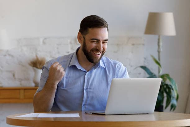 Man sitting at his laptop smiling and raising his fist in celebration of his success.